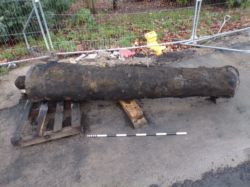 A large, black and rusted cannon lies on wooden pallets on paved ground, with a black and white measuring stick in front of it and a metal fence behind it.