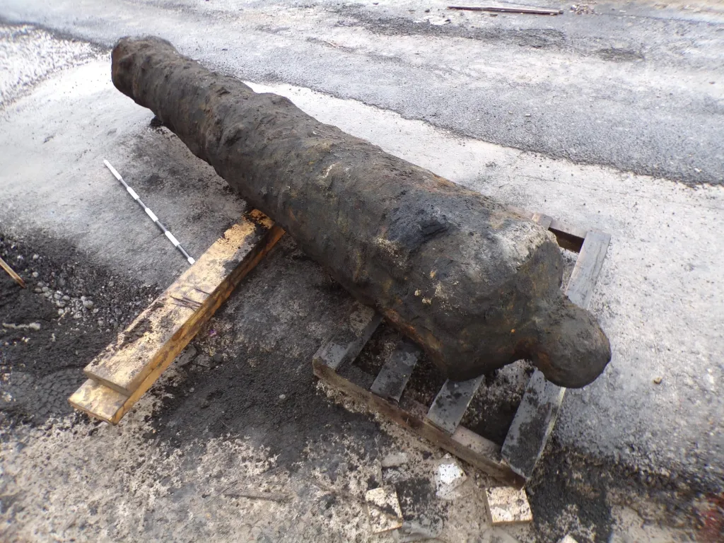 A large, dark, 300-year-old cannon resting on wooden pallets on an asphalt surface.