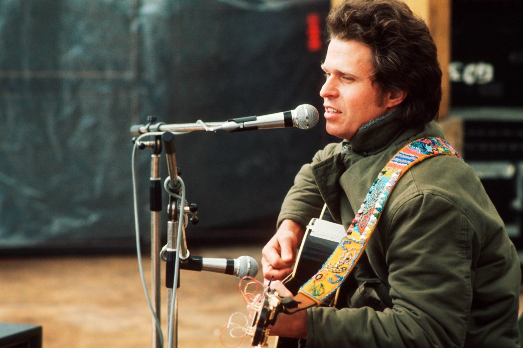Country Joe McDonald performing on stage, playing an acoustic guitar and singing into a microphone.