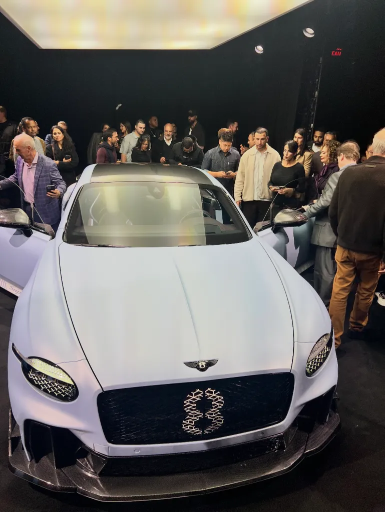 A light blue Bentley car with an "8" on the grill on display, surrounded by a crowd of people.