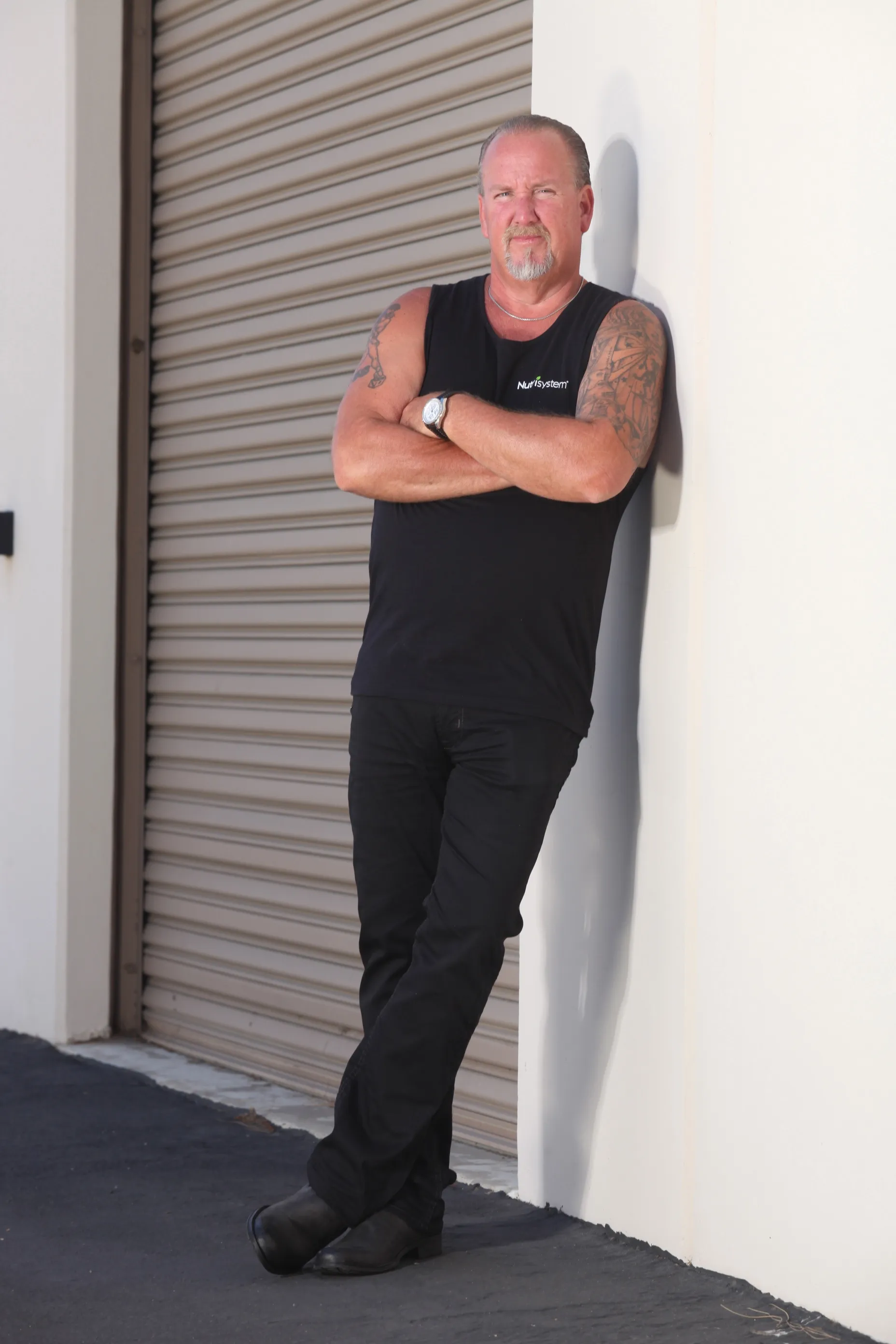 Darrell Sheets in a black tank top and jeans, leaning against a white wall with a closed garage door behind him.