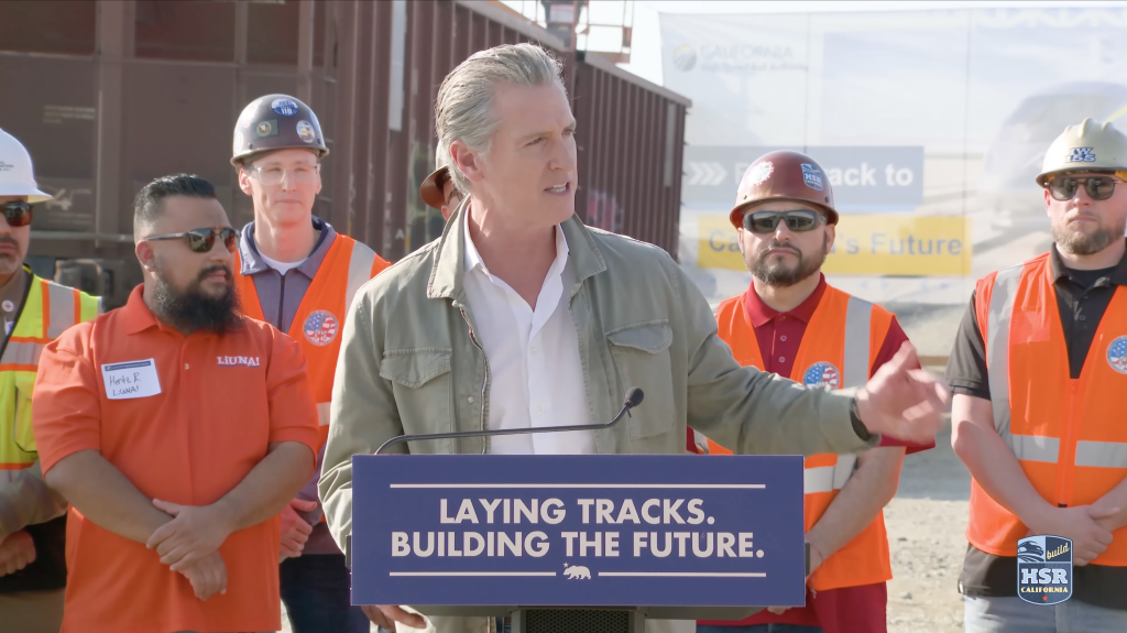 Gavin Newsom speaking at a podium with the text "LAYING TRACKS. BUILDING THE FUTURE." surrounded by construction workers.