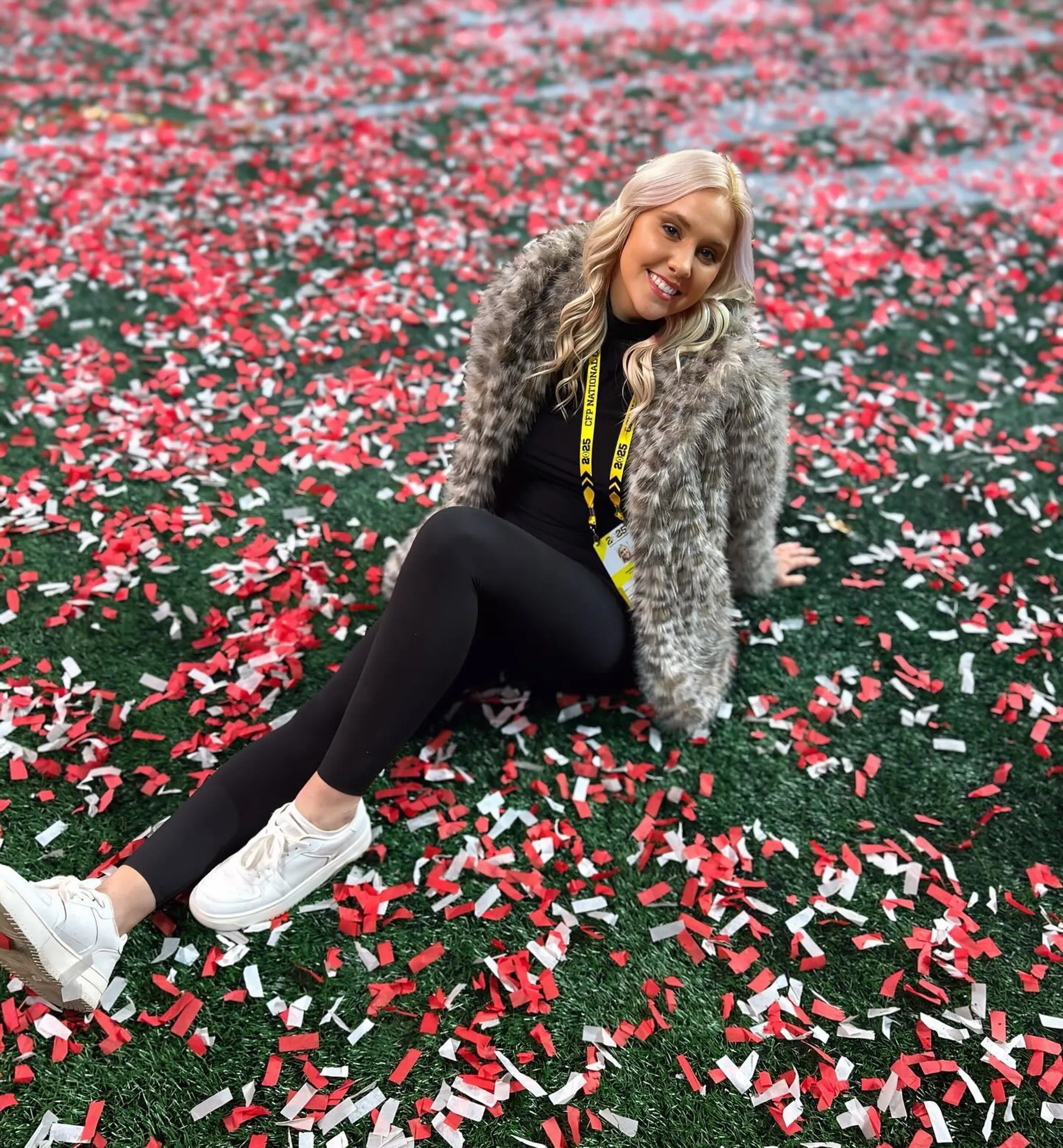A blonde woman wearing a furry coat sits on an astroturf field covered in red and white confetti.