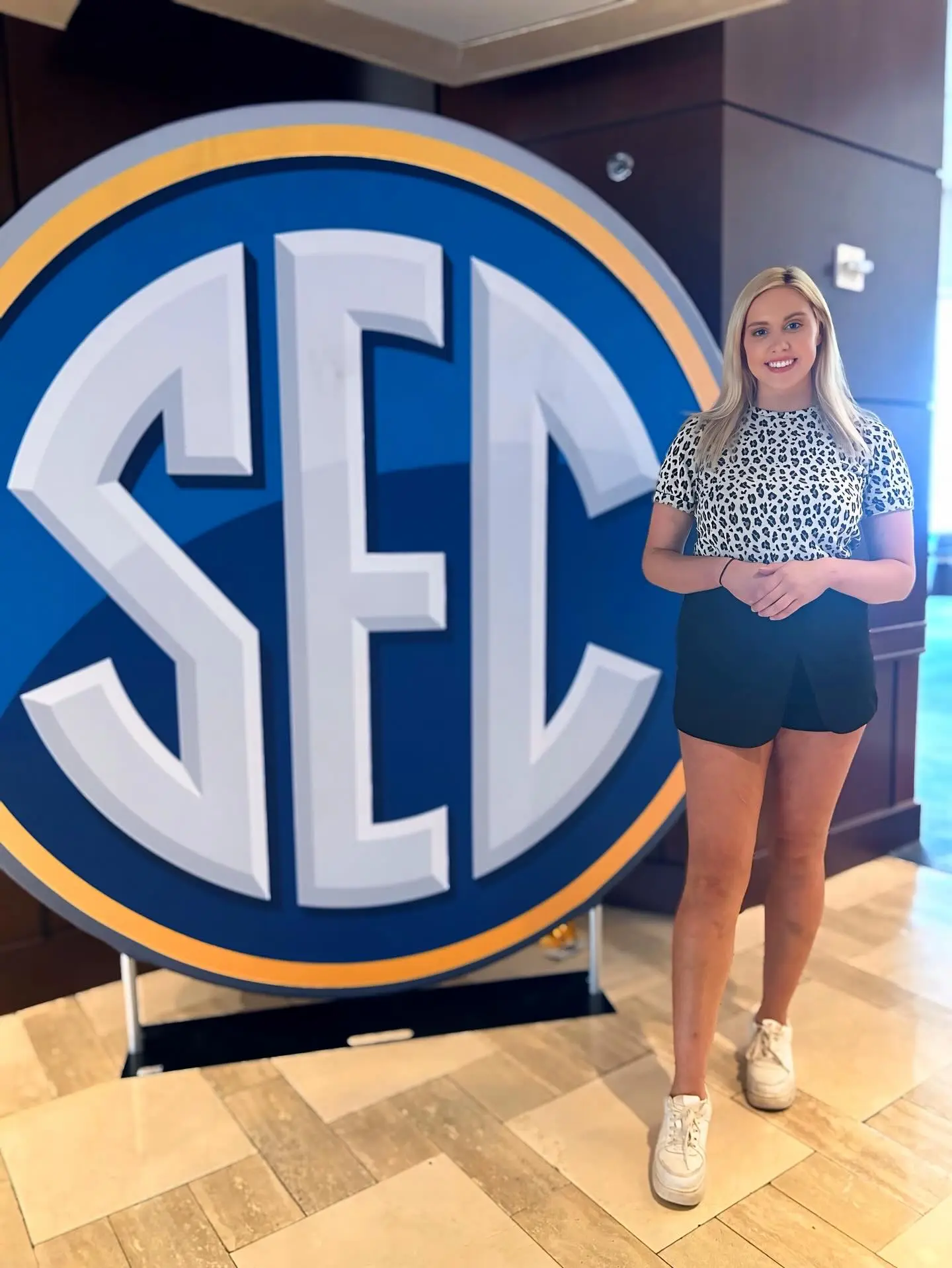 A woman in a leopard print top and black shorts stands next to a large SEC logo.