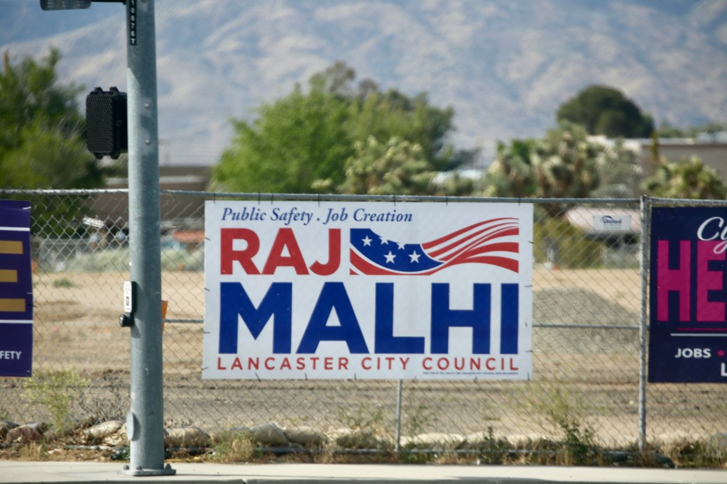 A "Raj Malhi Lancaster City Council" campaign sign with "Public Safety" and "Job Creation" on a chain-link fence.
