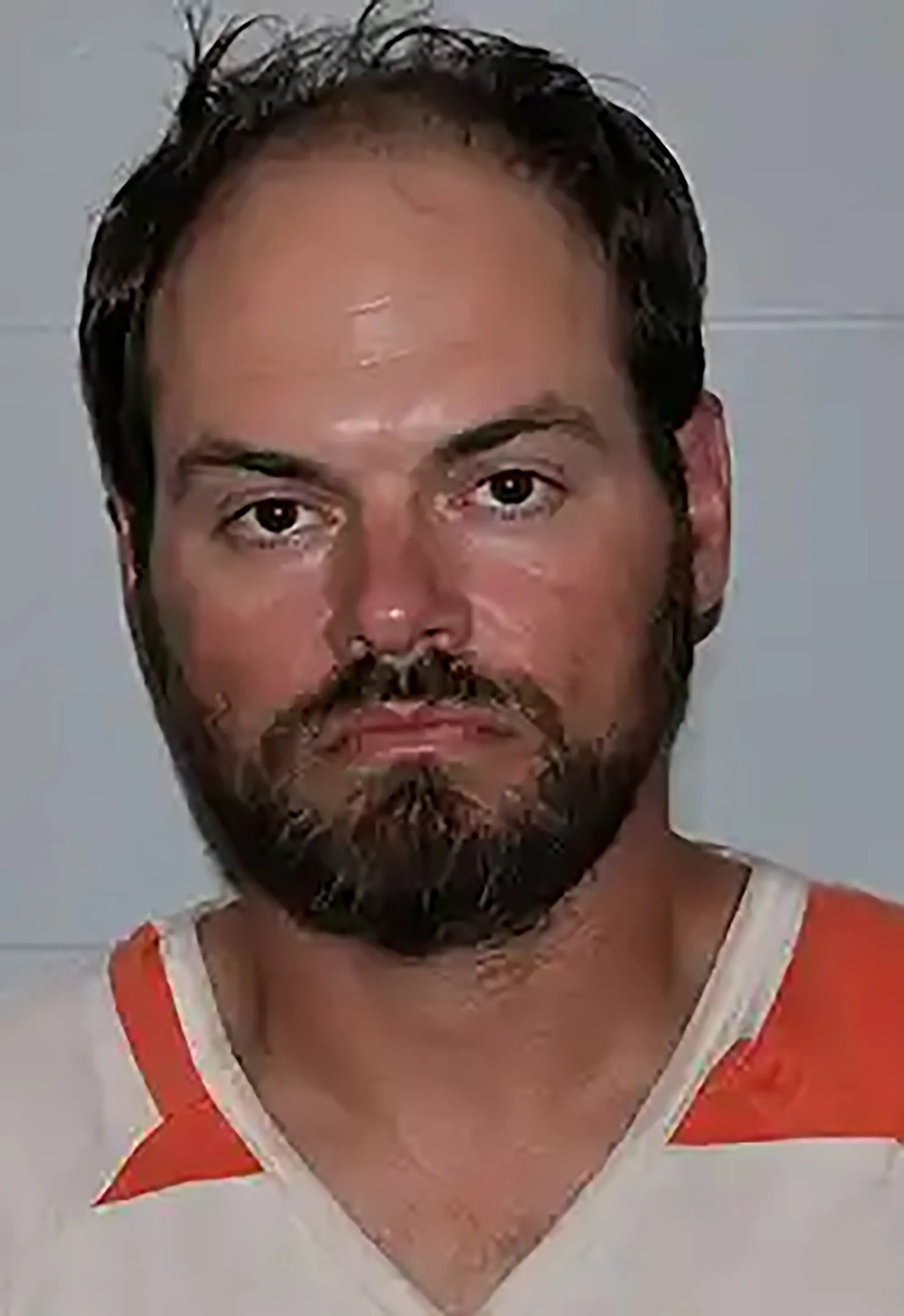 A close-up mugshot of a man with dark hair, a beard, and a mustache, wearing an orange and white striped shirt.
