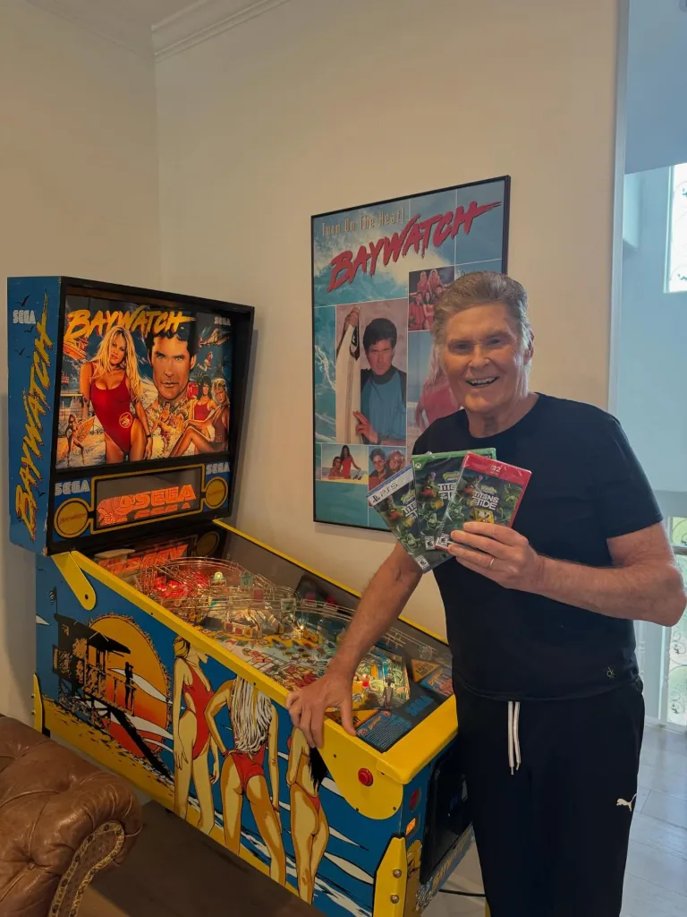 David Hasselhoff stands next to a Baywatch pinball machine and poster, holding three "Teenage Mutant Ninja Turtles" video games.