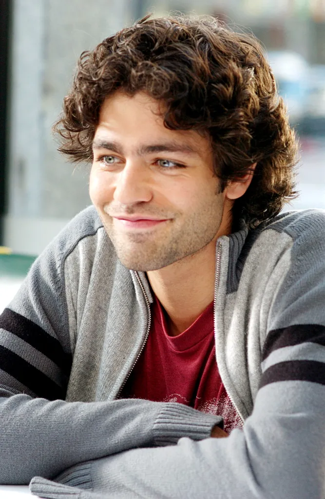 Adrian Grenier smiling in a gray striped jacket over a red T-shirt.