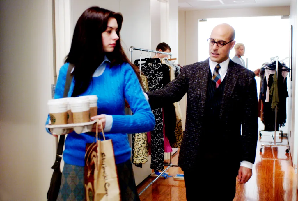 Anne Hathaway carrying a tray of Starbucks coffee, being guided by Stanley Tucci in a hallway with clothing racks.