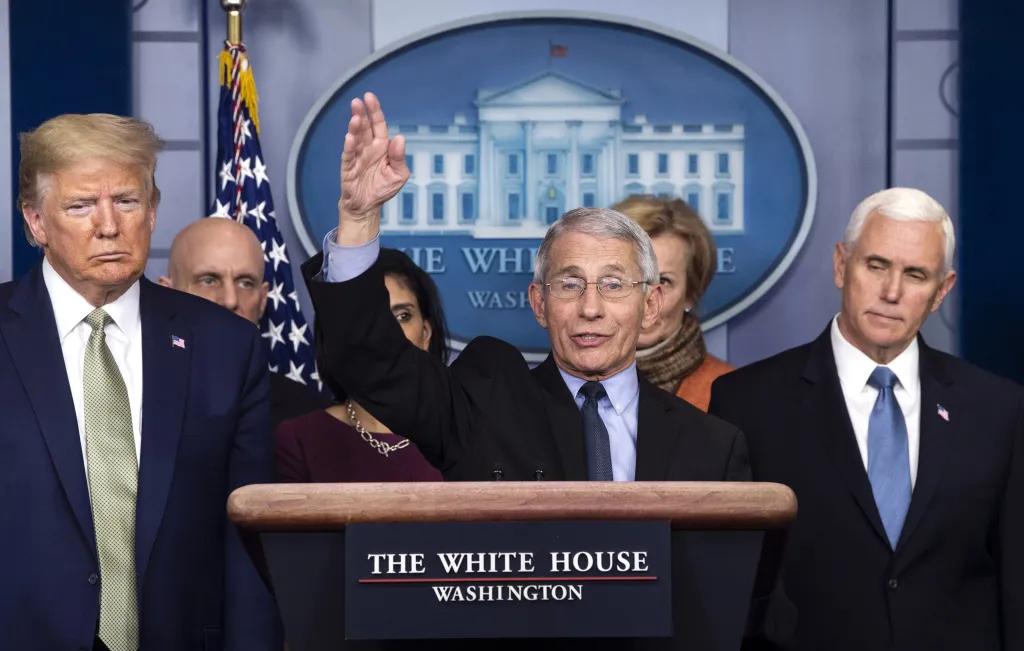 Dr. Anthony Fauci raising his hand to speak at a White House Coronavirus Task Force briefing with President Donald Trump and Vice President Mike Pence standing behind him.