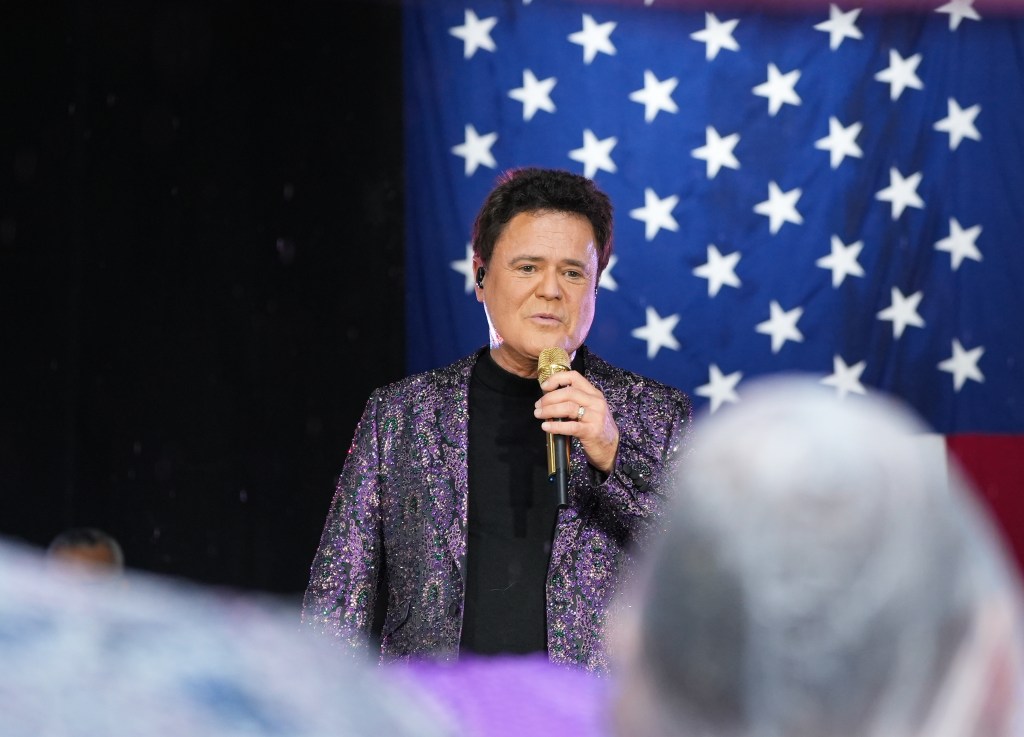Donny Osmond performing in a sequined jacket, holding a microphone, with an American flag in the background.