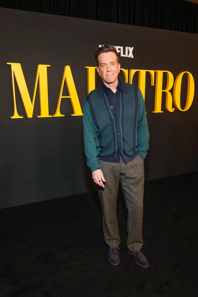 Ed Helms standing on a black carpet in front of a black wall with the yellow word "MAESTRO" on it.