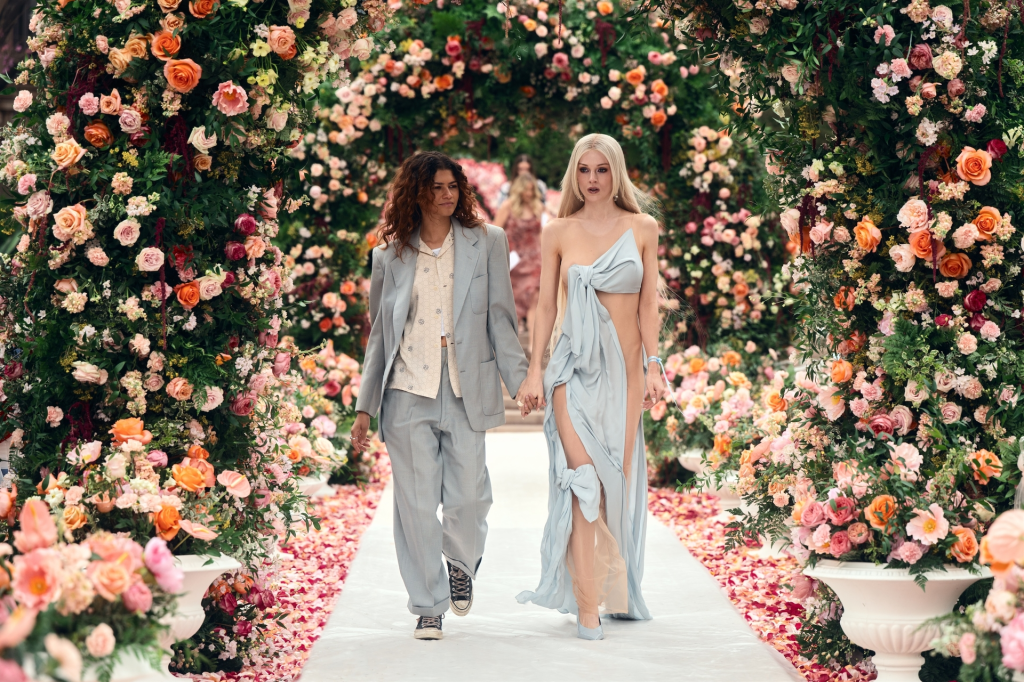 Zendaya and Hunter Schafer holding hands on a flower-lined runway in Euphoria.