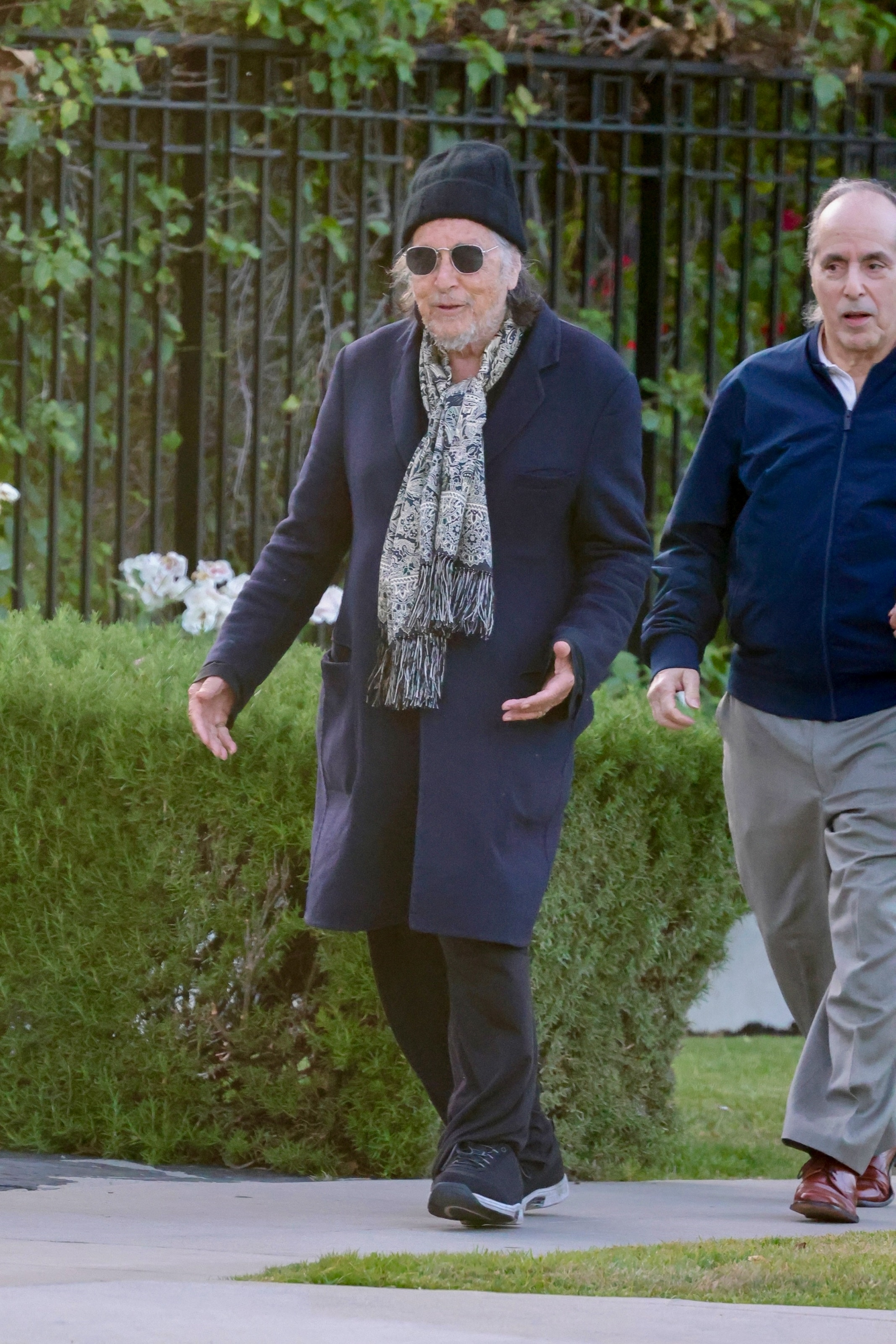 Al Pacino in a dark coat, scarf, beanie, and sunglasses walking outdoors with another man.