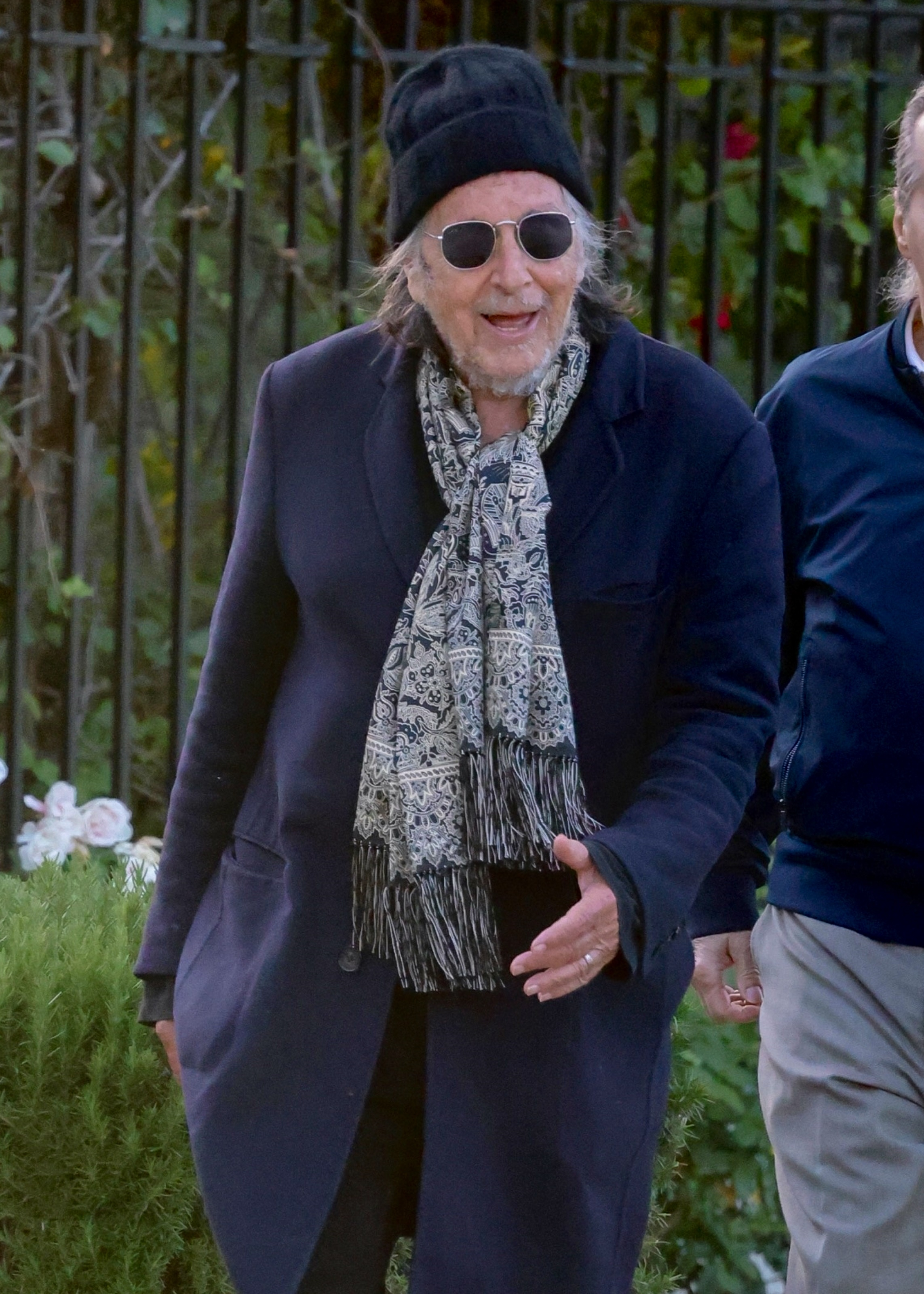 Al Pacino smiling, wearing a black beanie, sunglasses, a patterned scarf, and a dark coat during an evening stroll.