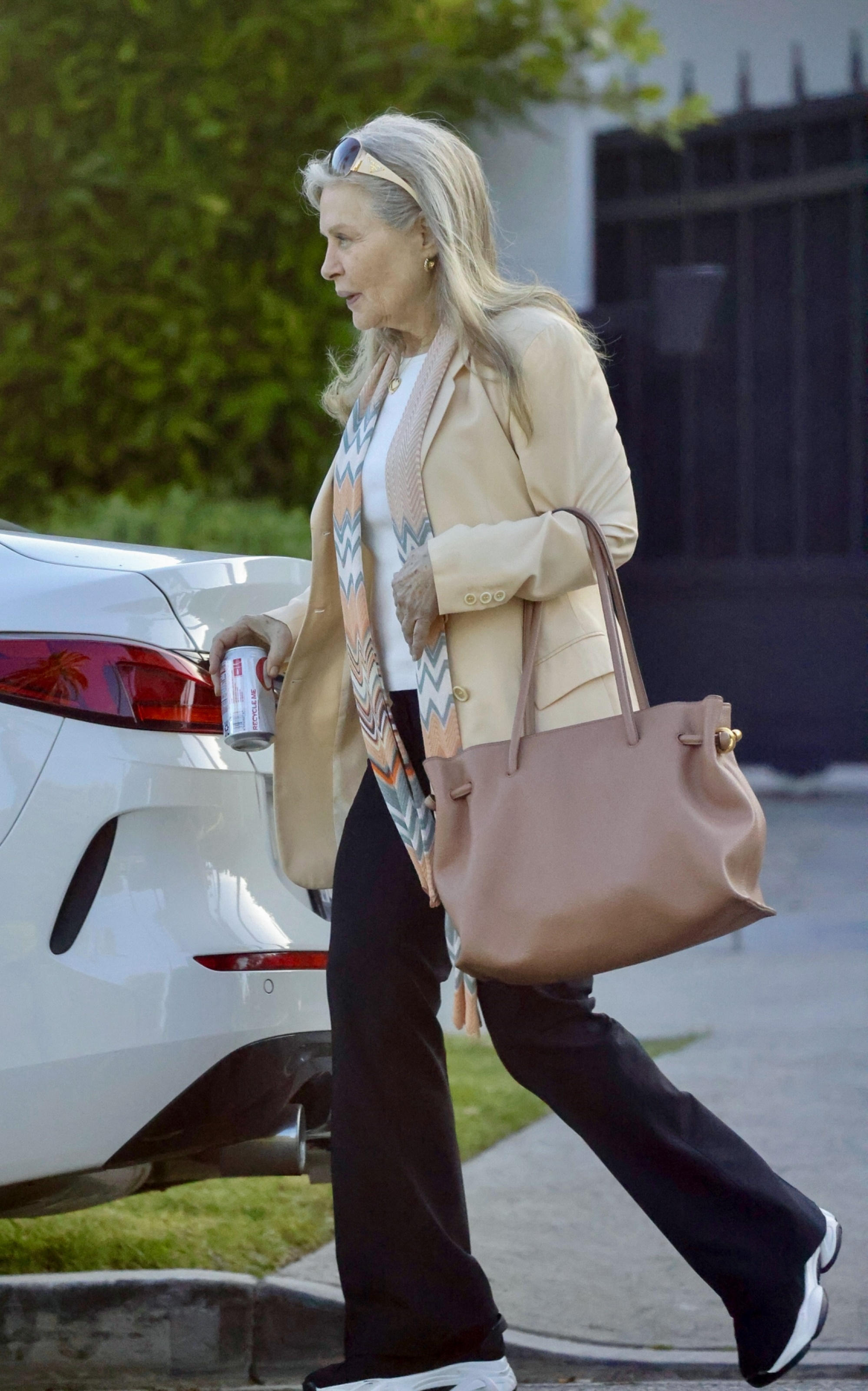 Beverly D'Angelo walking past a white car, holding a drink can, wearing a tan blazer, black pants, and a patterned scarf.