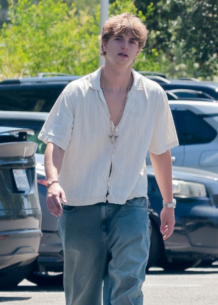 Jayden Federline wearing a light-colored, short-sleeved shirt, blue jeans, and a pendant necklace, walking in a parking lot.