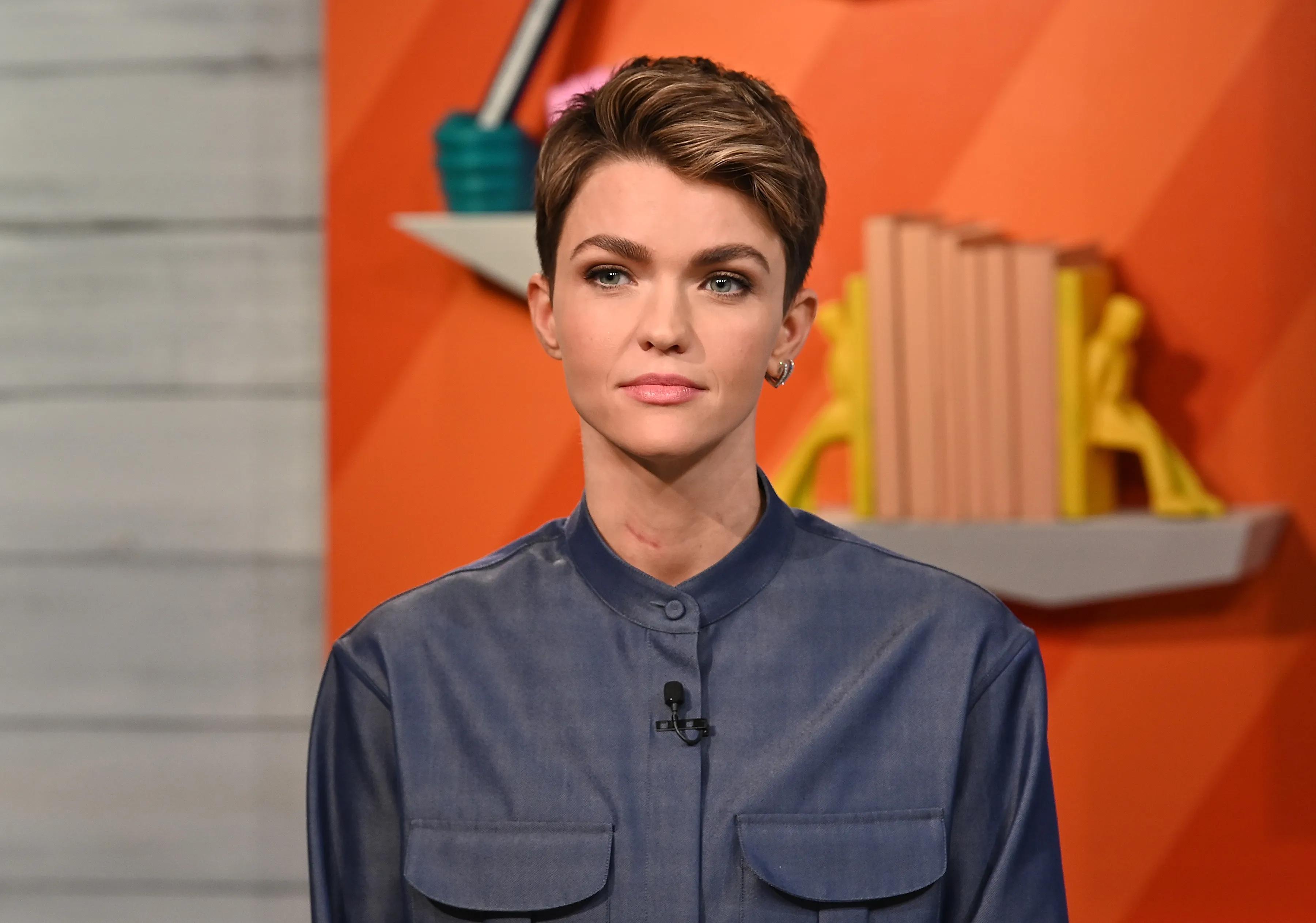 Ruby Rose wearing a blue collared shirt with two pockets on the chest, a small microphone clipped to the shirt, and a reddish mark on her neck.