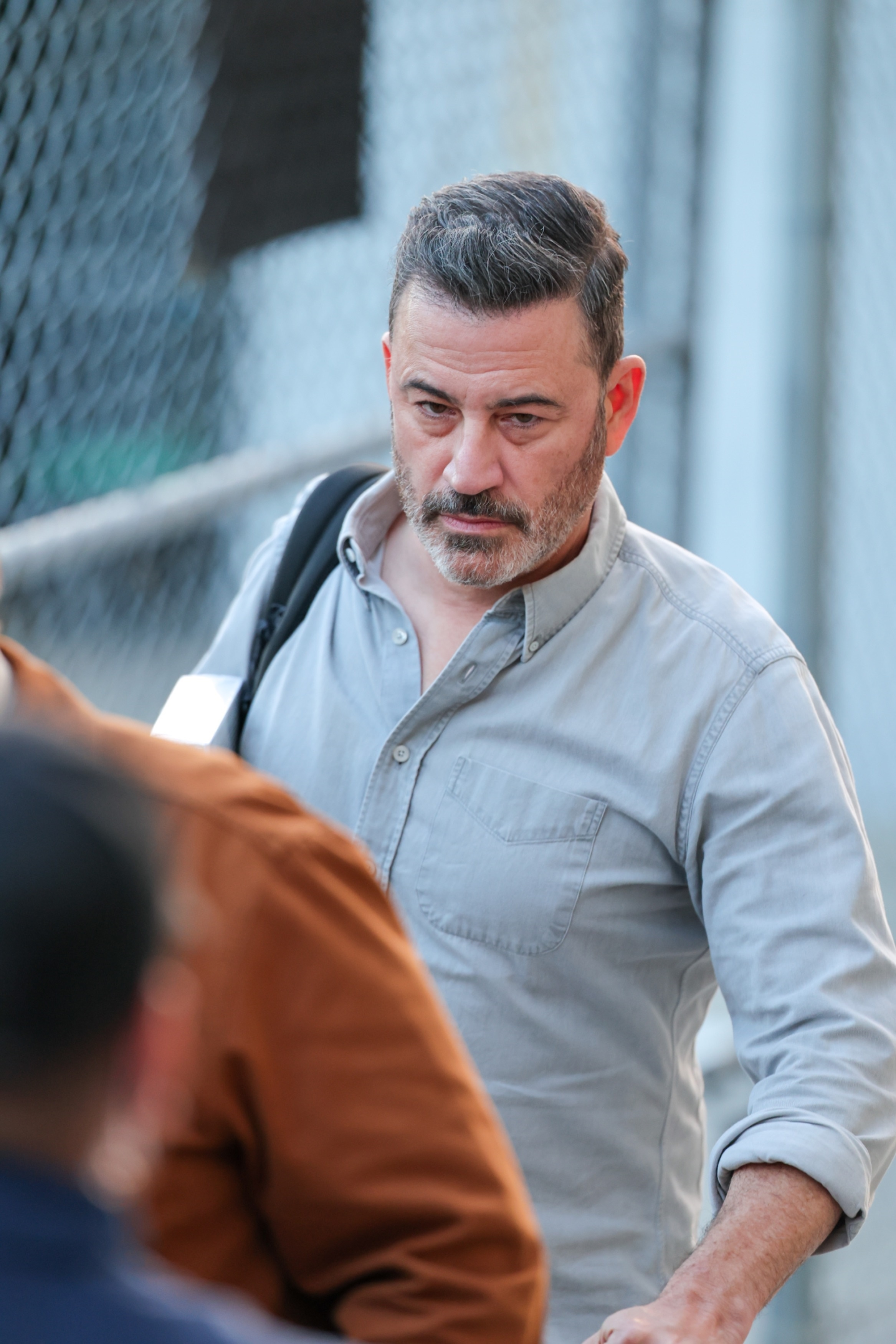 Jimmy Kimmel looking tense in a light gray button-up shirt and backpack.