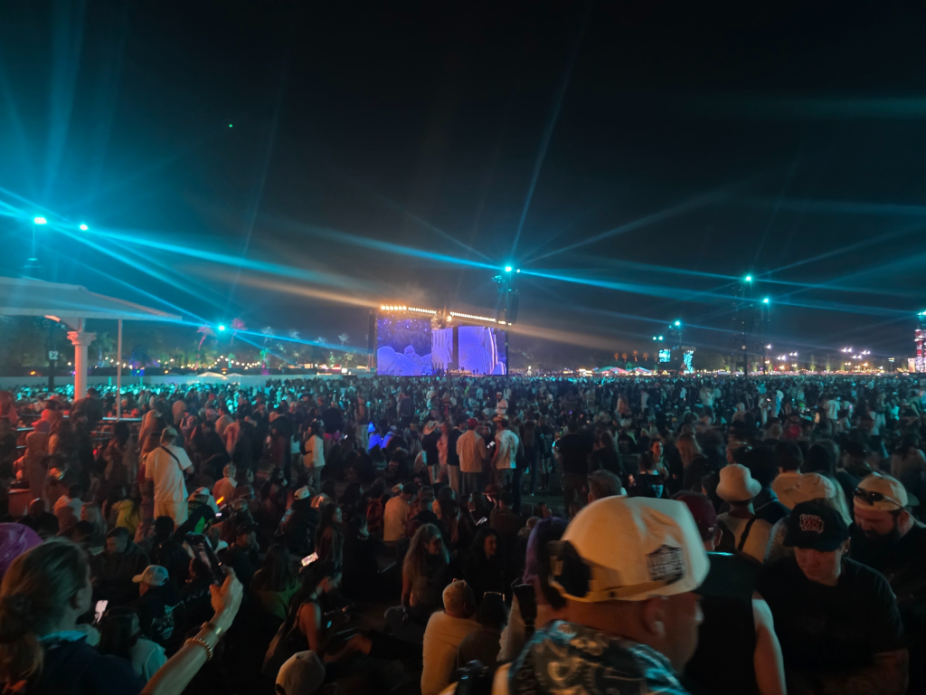 A crowd of fans gathered at the Coachella Music Festival, with bright blue lights shining across the night sky and a large stage visible in the distance.