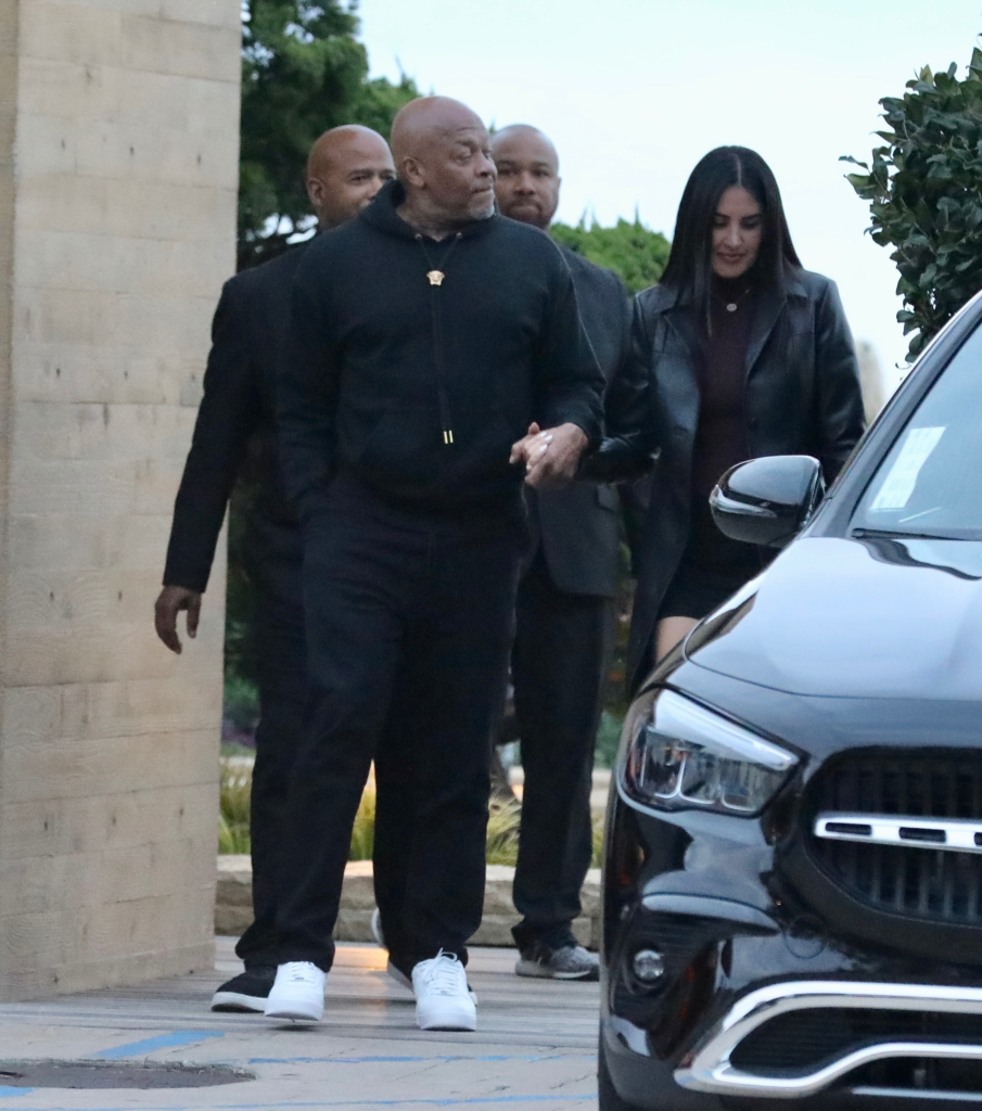 Dr. Dre in a black hoodie and black pants with two men and a woman in a black leather jacket next to a black car.