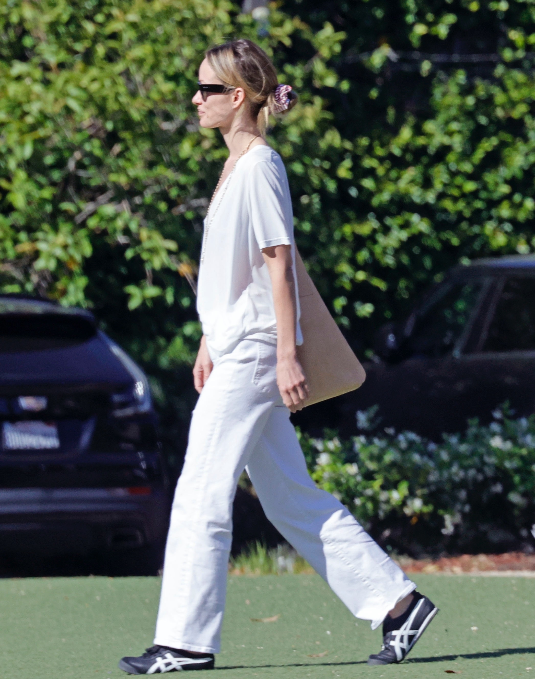 Olivia Wilde in a white outfit, walking in sunglasses with her hair in a bun.