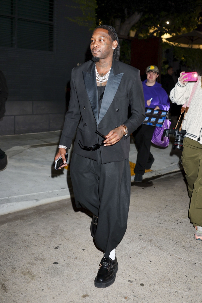 Offset wearing a black suit with a large diamond necklace, carrying sunglasses and a phone.