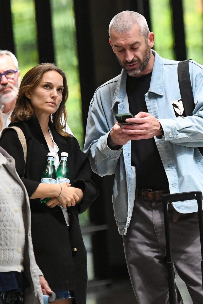 Natalie Portman holding two water bottles next to Tanguy Destable, who is looking at his phone, both seen at Montparnasse Station.