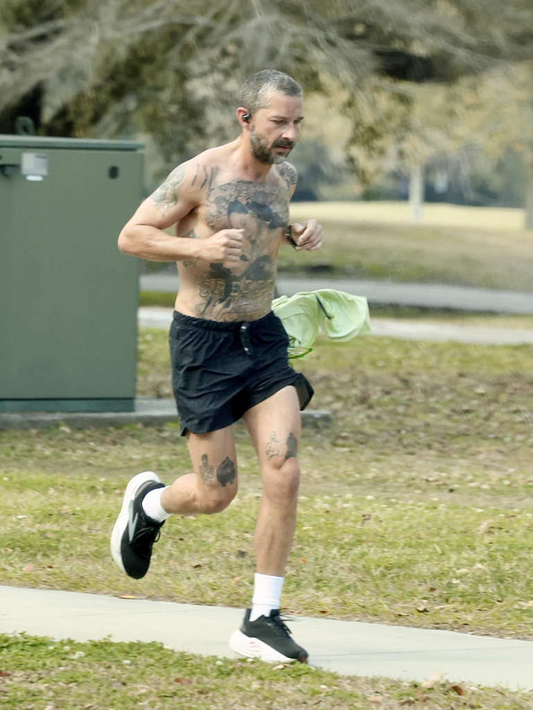 Shia LaBeouf jogging shirtless with numerous tattoos, wearing black shorts and sneakers.