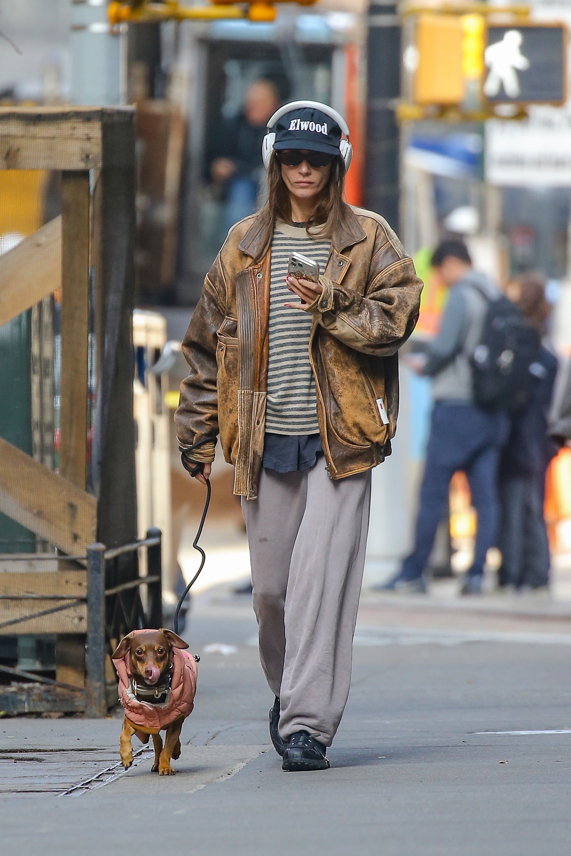 Amanda Batula walking her dog while wearing a brown leather jacket, striped shirt, grey sweatpants, sunglasses, and an Elwood baseball cap with headphones over it.