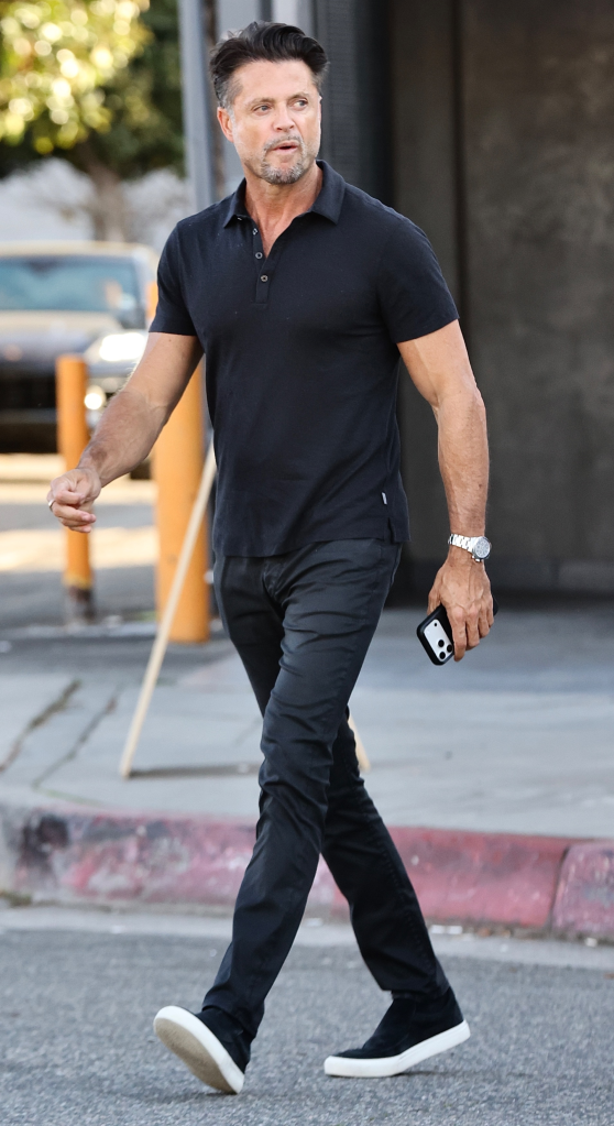 David Charvet walking in a black polo shirt, black trousers, and white-soled black sneakers, carrying a smartphone.