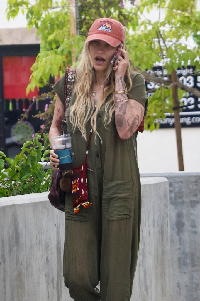 Paris Jackson walking and talking on a phone, holding a cold drink, wearing a green jumpsuit and a baseball cap.