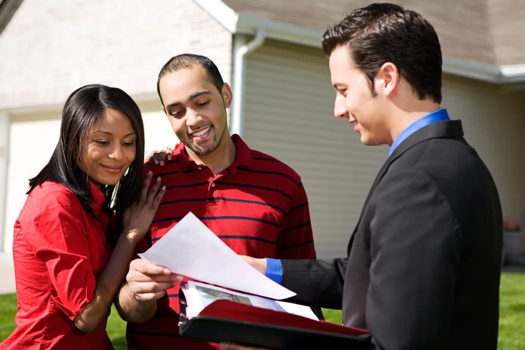 A real estate agent presenting a brochure to a smiling African American couple in front of a house.