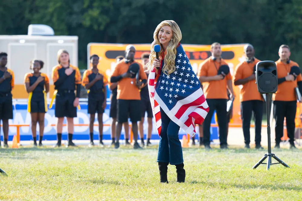 Heidi Gardner as Tisha Basmati, wrapped in an American flag, holding a microphone, with a crowd of people in the background.