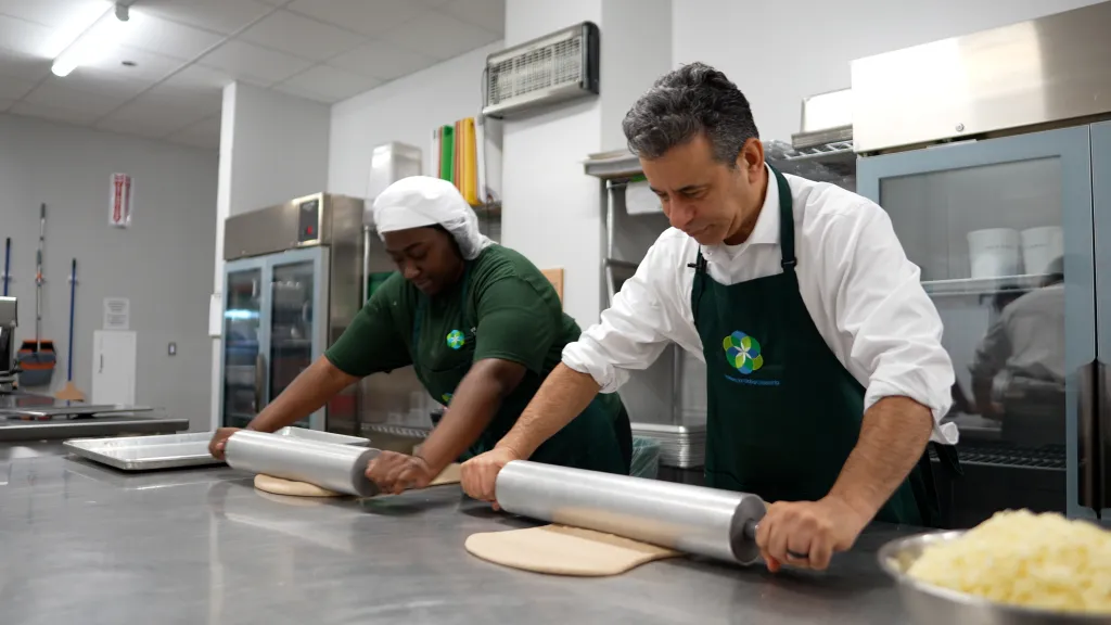 Two people roll out dough in a professional kitchen.