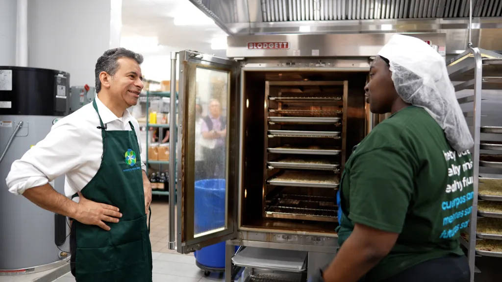 Two people in a kitchen, one wearing an apron, are looking at food trays in an open oven.