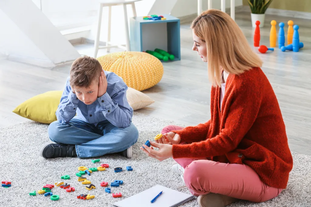 A female psychologist using letter blocks to work with an autistic boy who is covering his ears and looking upset.