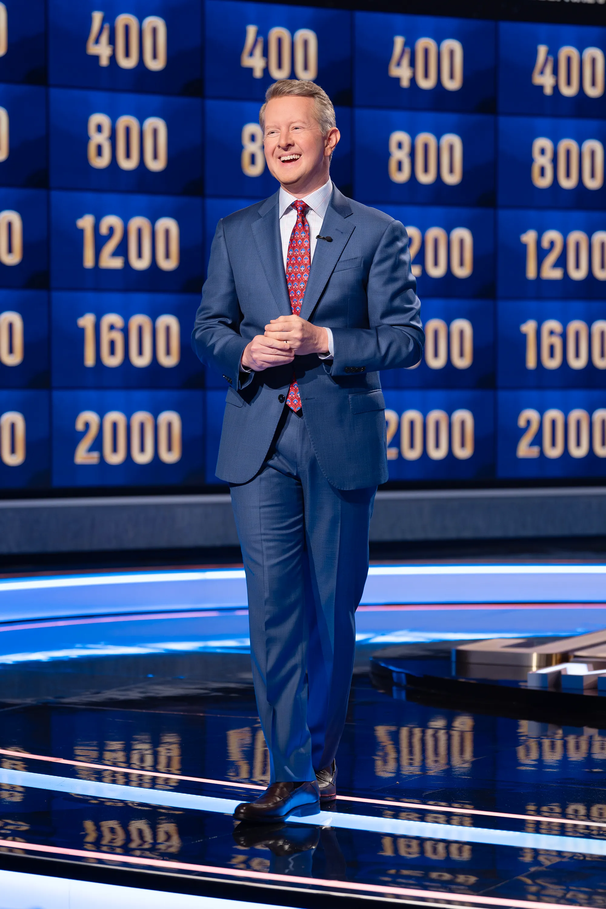 Ken Jennings standing on the "Jeopardy!" set with a blue backdrop displaying various point values.
