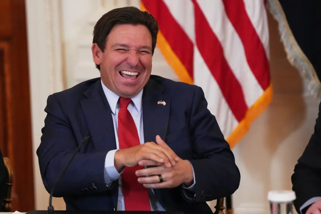 Florida Gov. Ron DeSantis laughing in front of an American flag during a roundtable discussion.
