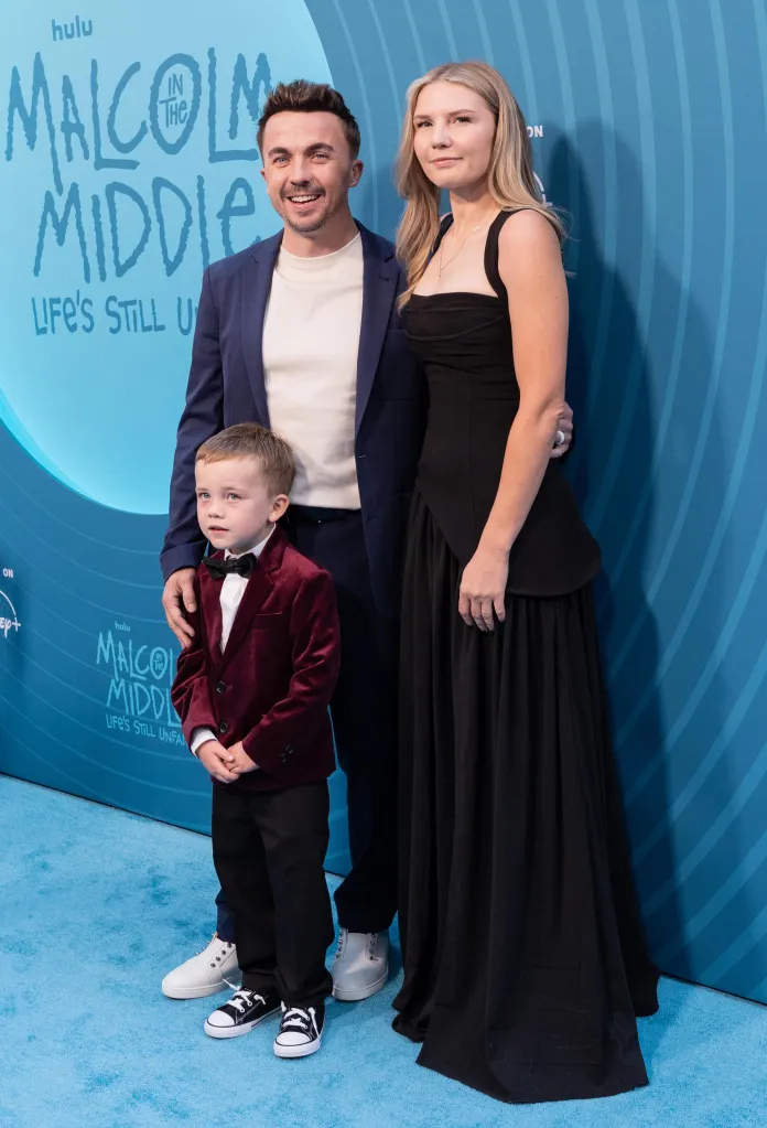 Frankie Muniz, his wife, and son stand on a blue carpet in front of a "Malcolm in the Middle" banner at a Hulu event.
