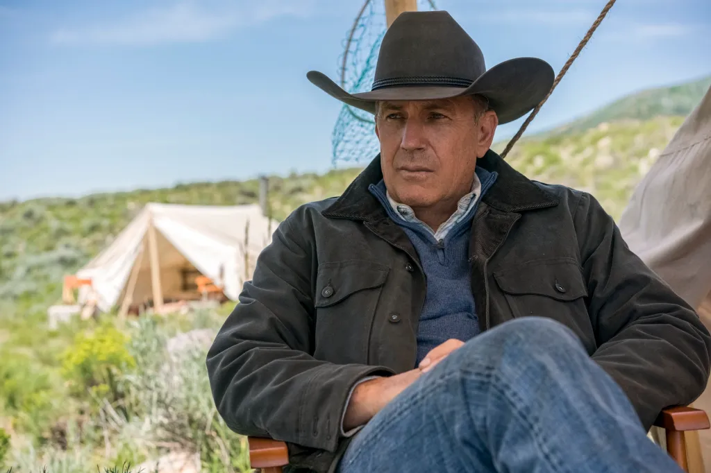 Kevin Costner as John Dutton in a cowboy hat and jacket, sitting outdoors.