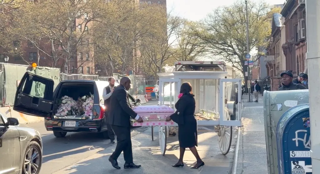 A man and woman carrying a pink casket for a baby, alongside a white horse-drawn carriage and a hearse filled with flowers, on a city street.