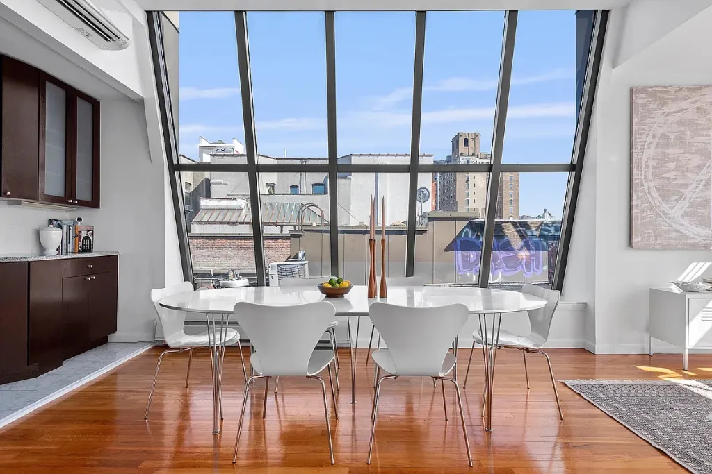 A modern dining room with a white table and chairs, a brown kitchen counter with cabinets, and large slanted windows overlooking city rooftops.