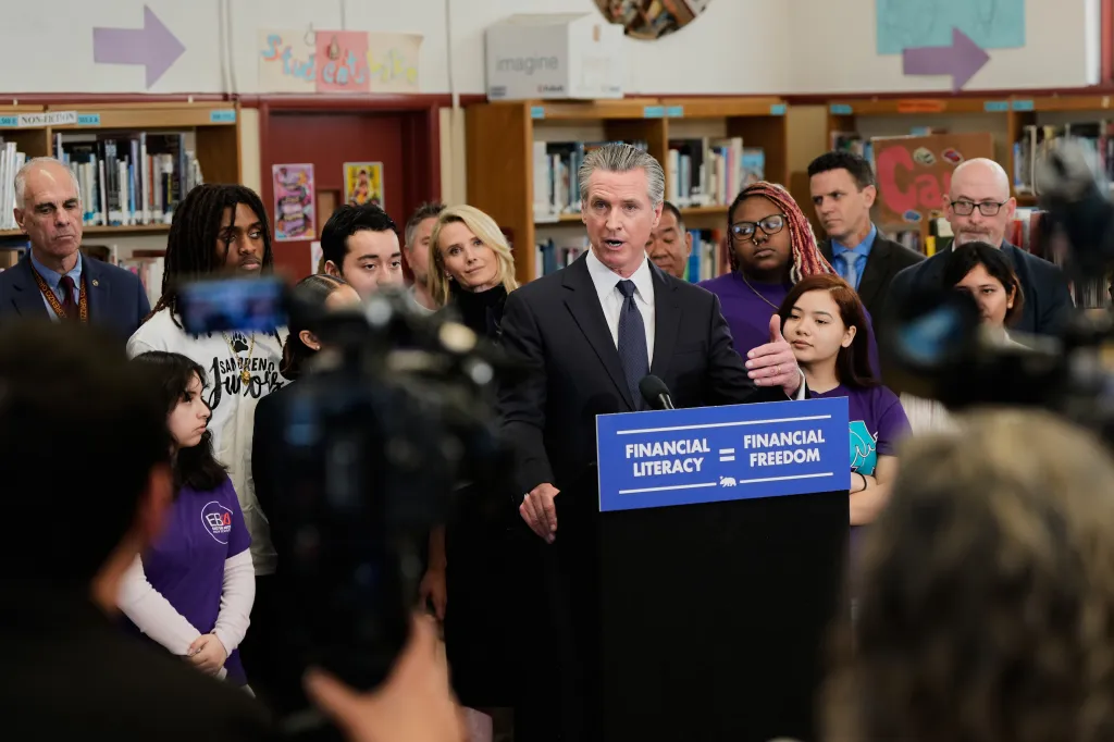 Governor Gavin Newsom speaks at a press conference in a school library, with a sign reading "Financial Literacy = Financial Freedom" on the podium.