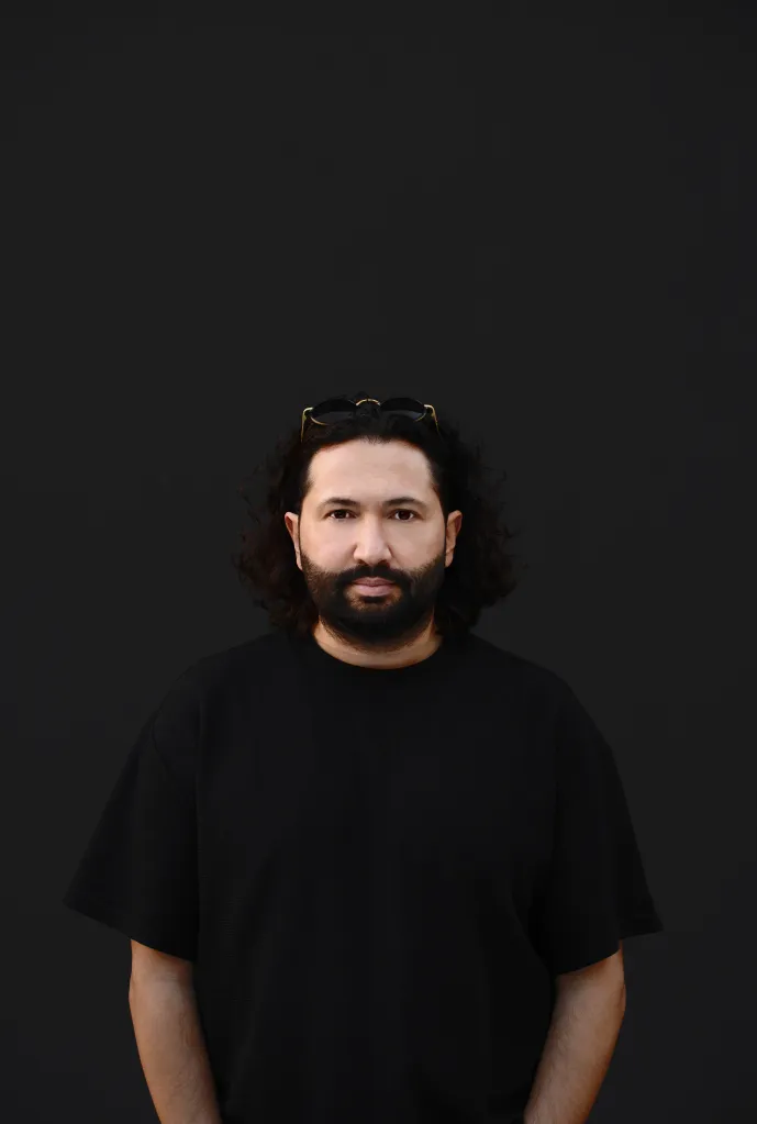 Photo of Ninetoes, a man with dark curly hair, a beard, and sunglasses resting on his head, wearing a black shirt.