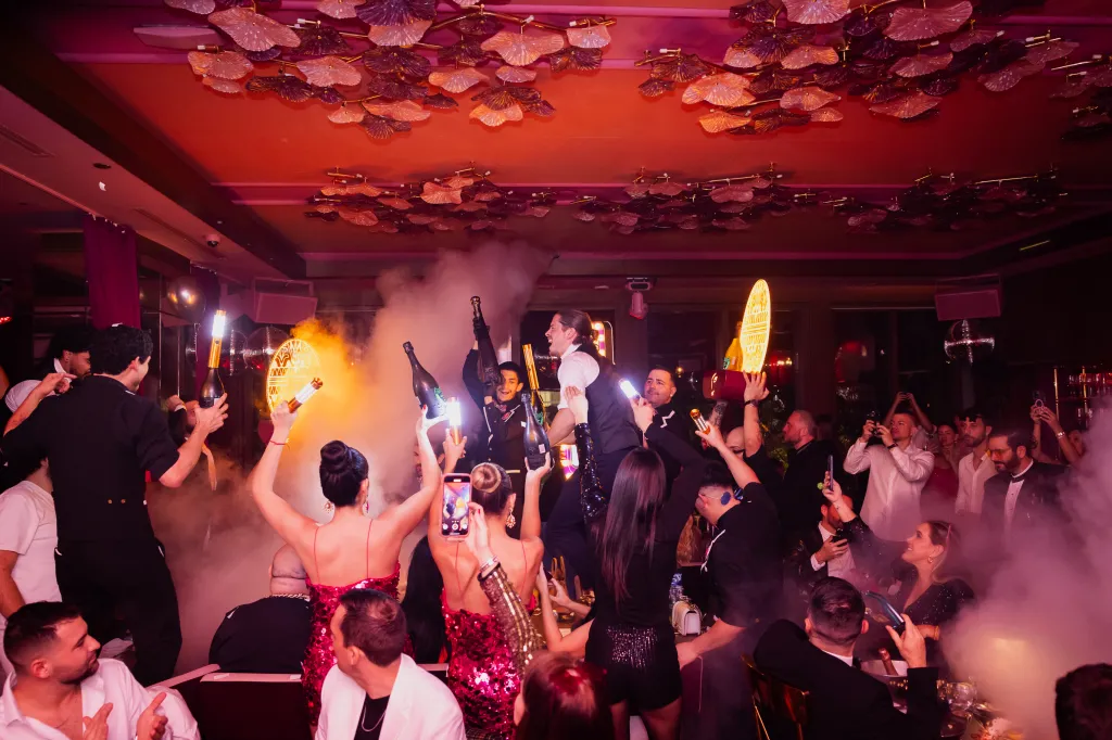 People raising champagne bottles and holding phones in a club with fog.