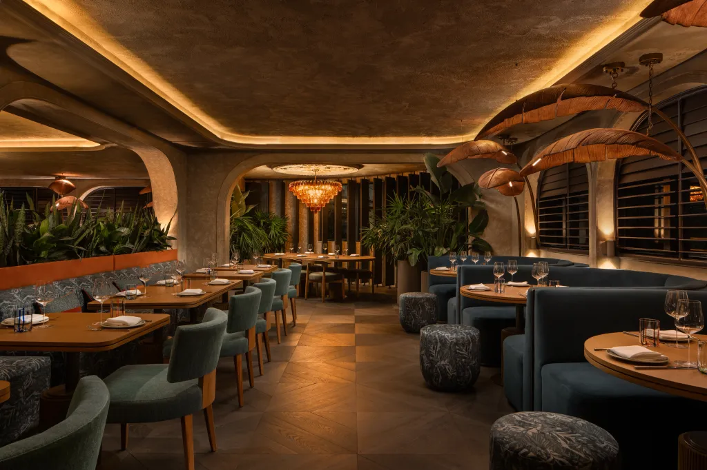 A modern dining room with blue and green seating, amber lighting, and tropical plants.