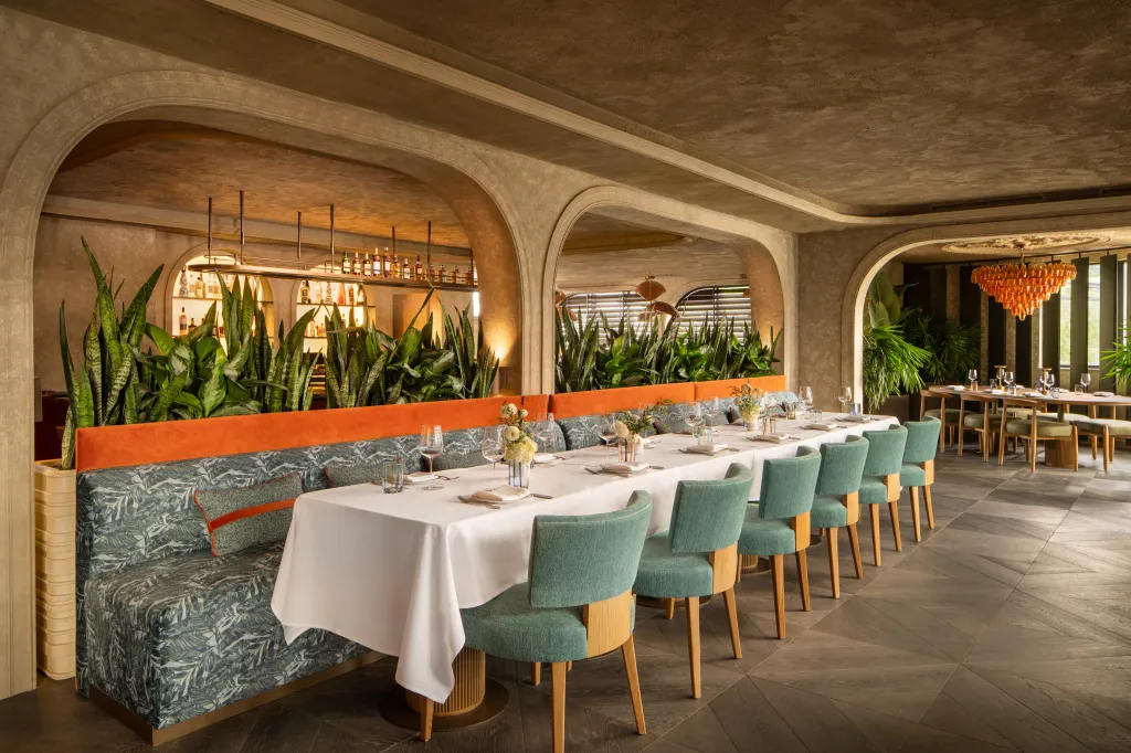 Long dining table set for guests with teal chairs and a botanical print banquette, with a full bar and plants in the background.