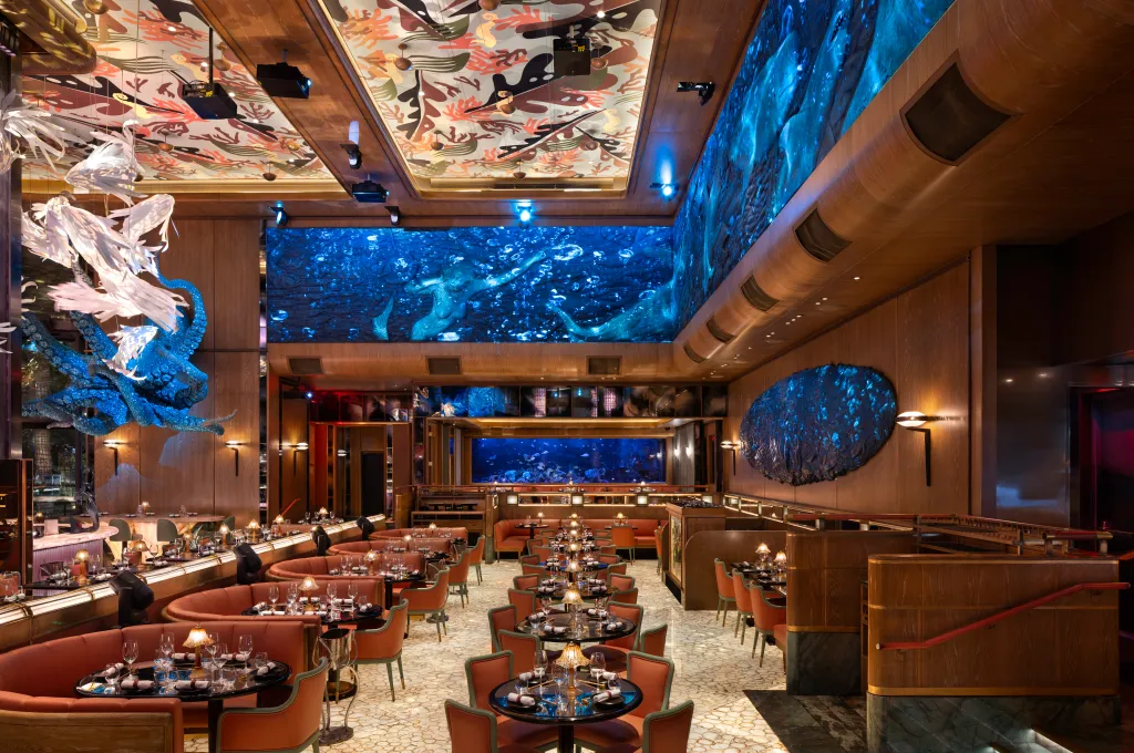 The interior of Sexy Fish restaurant, featuring a large screen displaying mermaids, an aquarium with fish, and coral-patterned ceiling art.