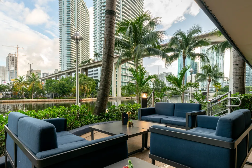 Outdoor seating area with blue couches and tables overlooking a canal with city buildings and palm trees in the background.